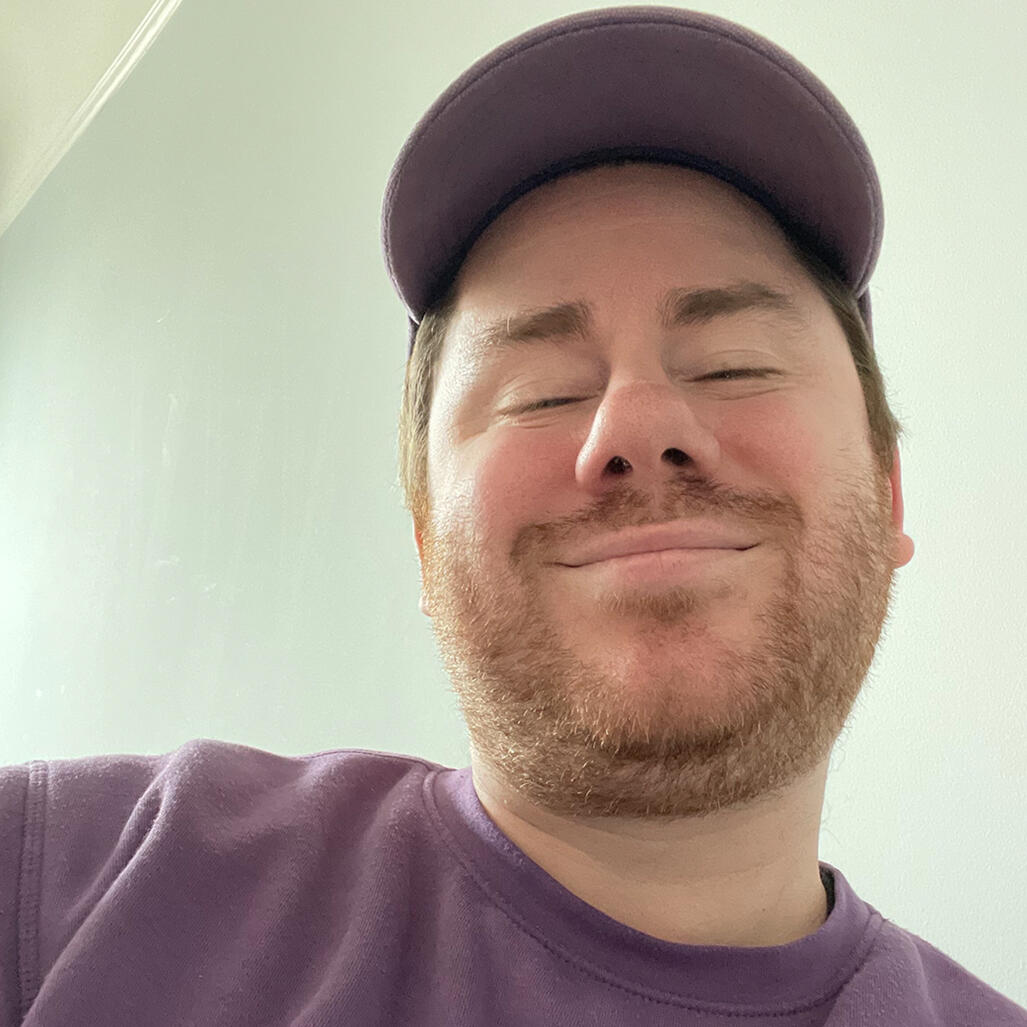 A photo of Mikey in a purple sweater and purple ballcap, smiling with his eyes closed.